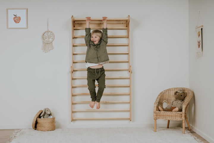 A child hanging from a wooden Pikler Triangle Swedish Wall indoors, with a basket and a wicker chair in the background.